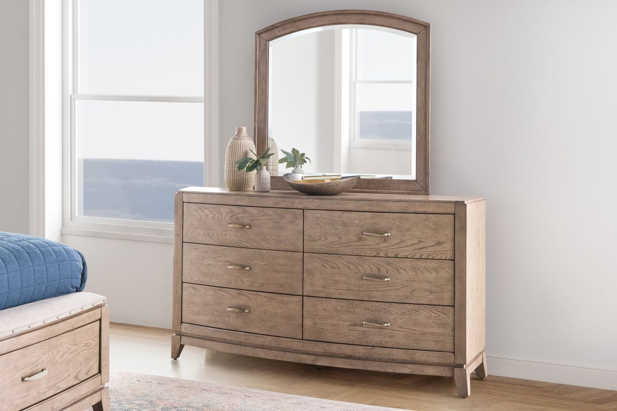 A calm, sunlit bedroom with a wood dresser and a well-proportioned rectangular mirror centered above it, styled with a small ceramic lamp and a simple vase