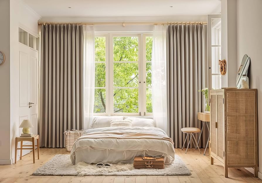 A cozy bedroom with blackout curtains hung high above a window, the panels falling to the floor beside a neatly made bed