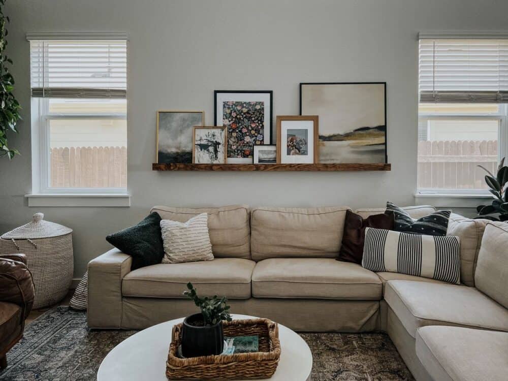 A living room sofa with a long picture ledge mounted above it, holding layered framed art and a small ceramic vase