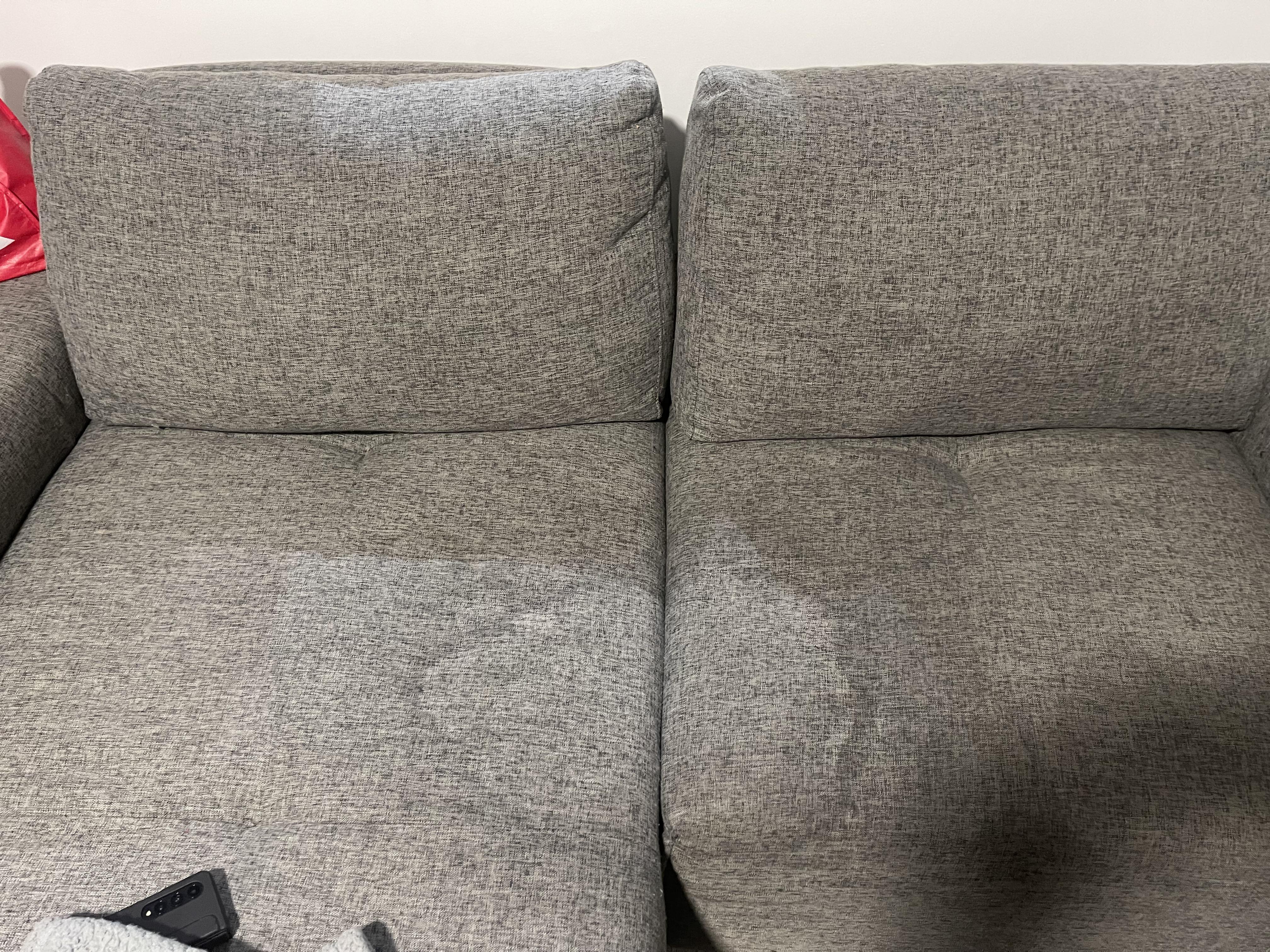 A person lightly misting rubbing alcohol onto a microfiber couch armrest while blotting with a clean white cloth