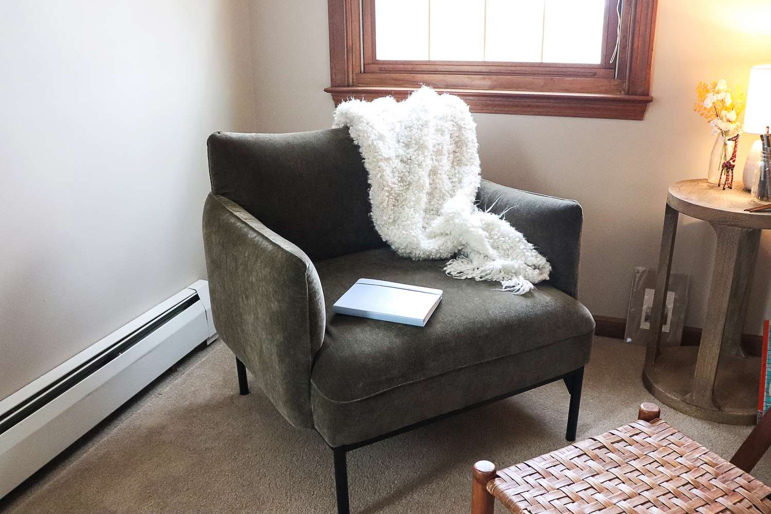 A small reading nook with a compact accent chair, a footstool, and a slim side table arranged in a tight apartment corner with warm lighting