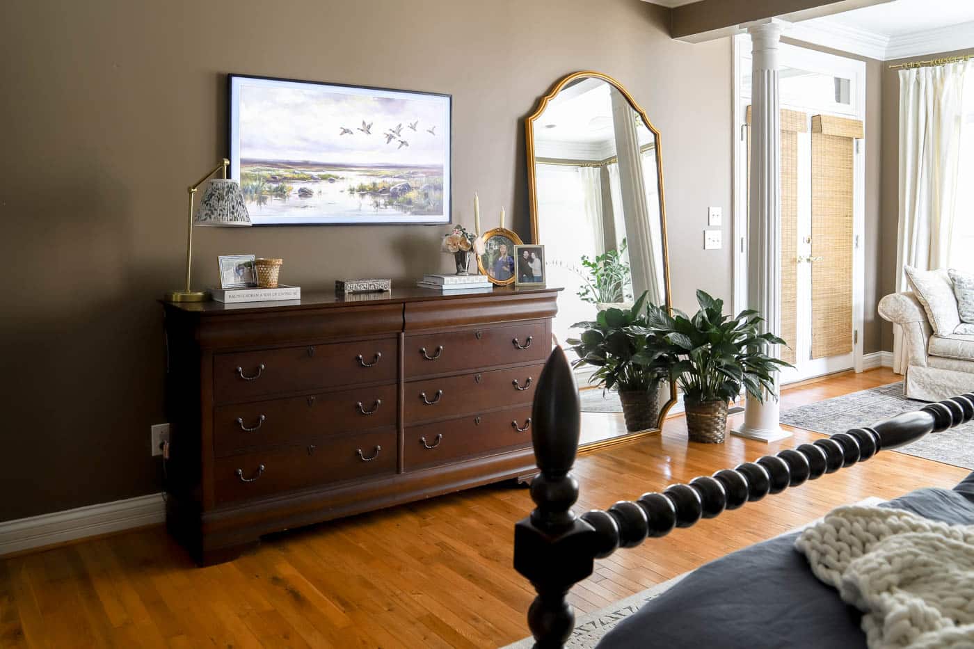 A tall framed mirror leaning securely against a wall on top of a dresser, with a small tray and a table lamp creating a relaxed layered look
